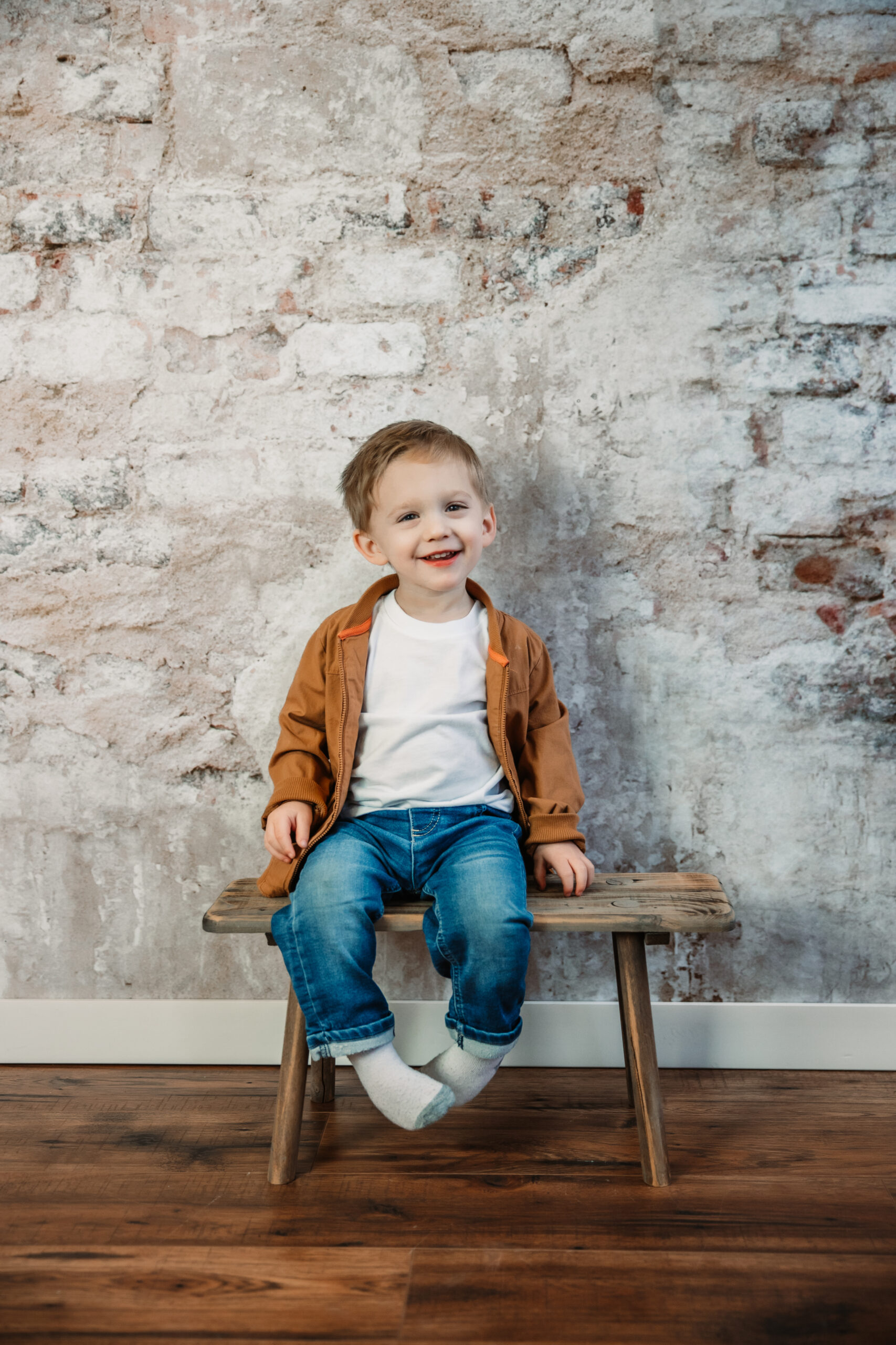 adorable two year old boy sits on bench in front of brick wall